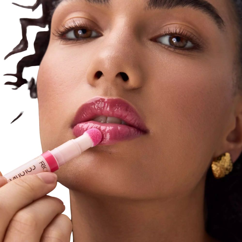 Persoon houdt een lipolie-applicator vast. Lippen bedekt met glanzende lipolie. Opschrift: COLOUR.