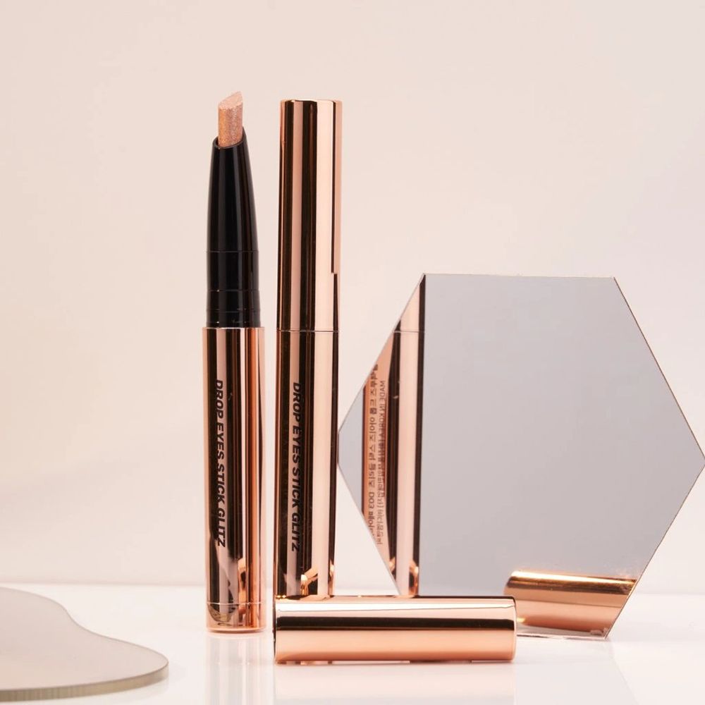 Crayons fard à paupières Drop Eyes Stick Glitz et miroir. Deux crayons, un ouvert. Emballage doré rose. Miroir en arrière-plan.
