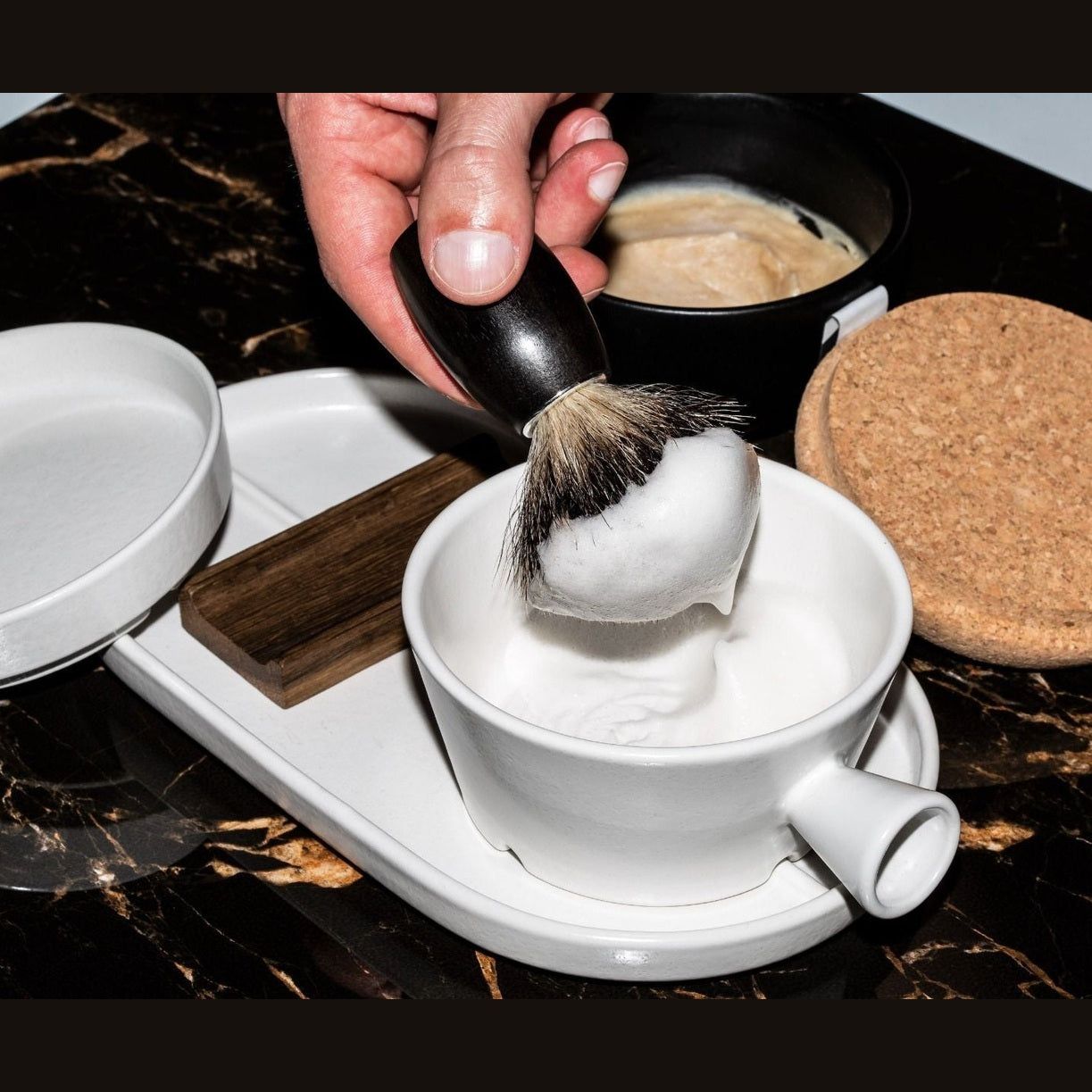 Scheerkwast met schuim in een kom. Andere items: zeepbakje, houten blok, kurken onderzetter en kom met scheercrème.