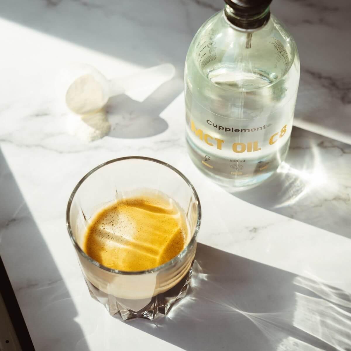 Glas met drank, daarnaast fles met etiket "Cupplement MCT OIL C8". Lepel op marmeren oppervlak.