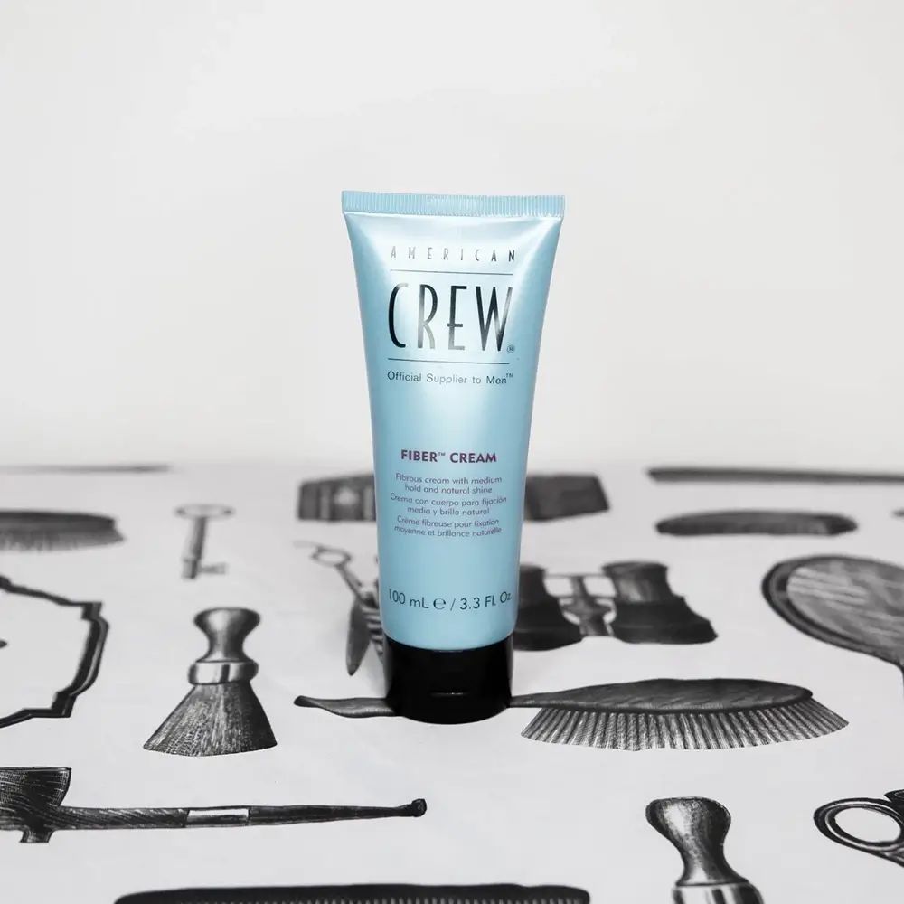 Lichtblauwe tube met zwarte dop. Opschrift: American Crew, Fiber Cream. Op tafel met kappersmotieven.