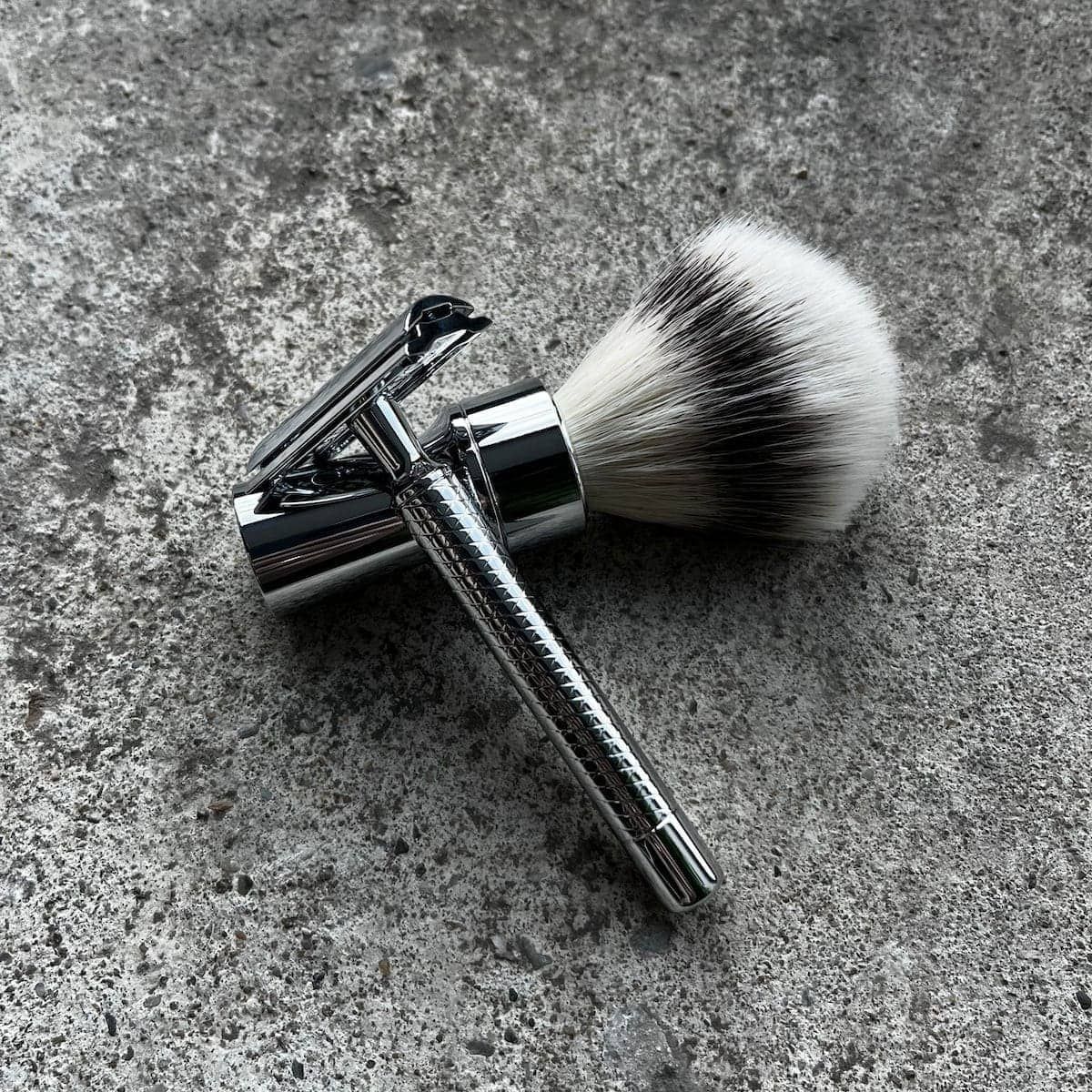 Blaireau à poils blancs et noirs et rasoir en métal. Les deux sont posés sur une surface grise.