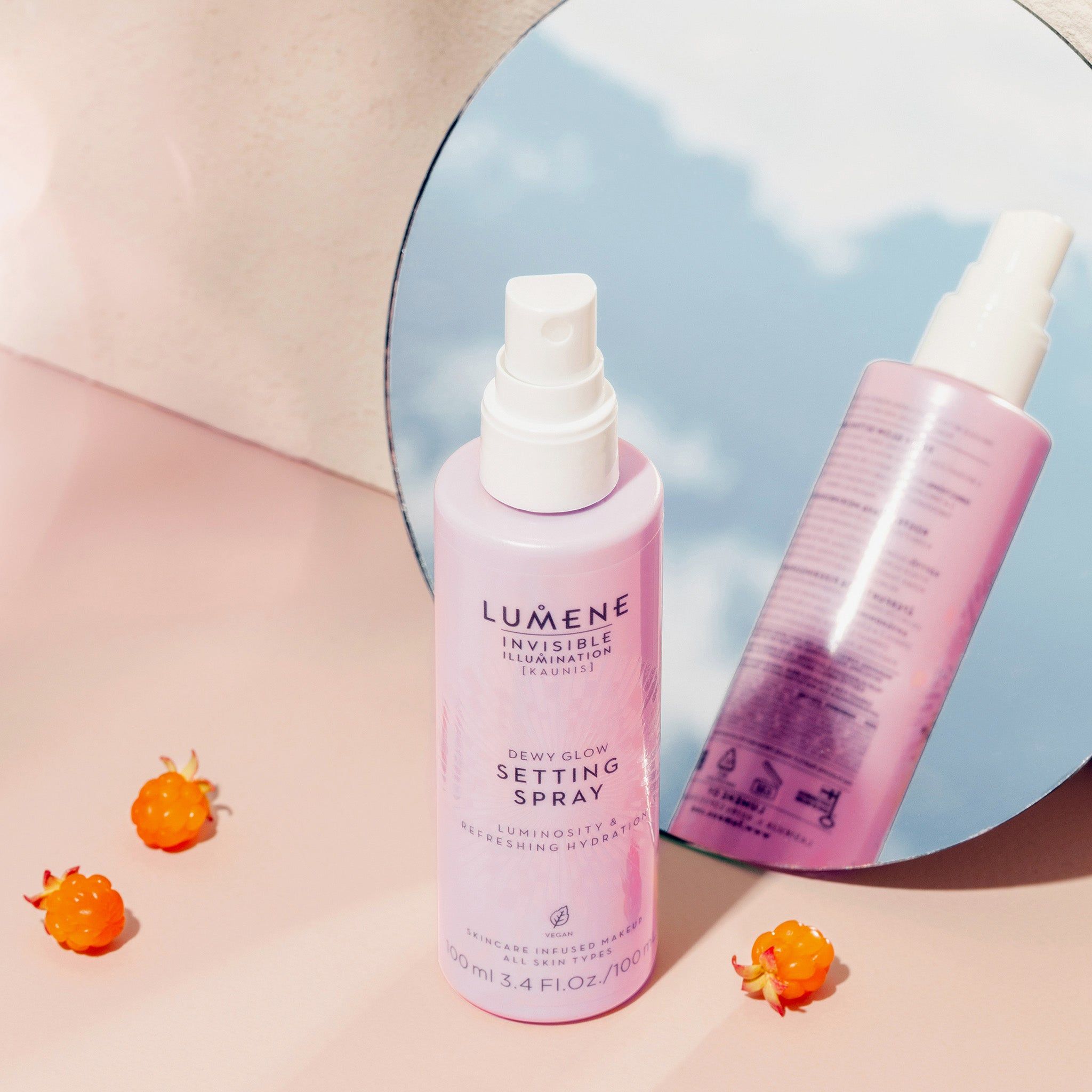 Twee roze flessen met witte spraykop. Opschrift: Lumene INVISIBLE ILLUMINATION Dewy Glow Setting Spray. Weerspiegeling in spiegel. Bessen.