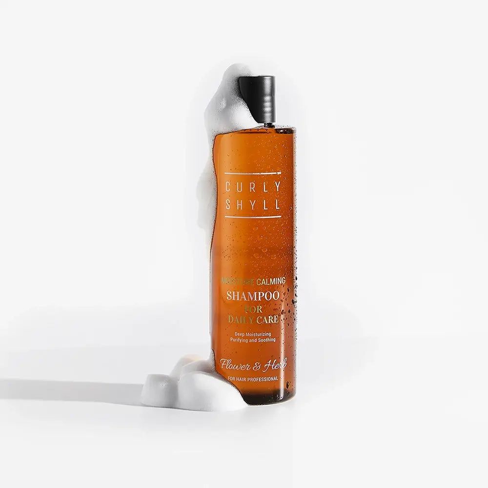 Bouteille marron avec mousse. Inscription: Curly Shyll, Moisture Calming Shampoo. Marque: Flower & Herb.