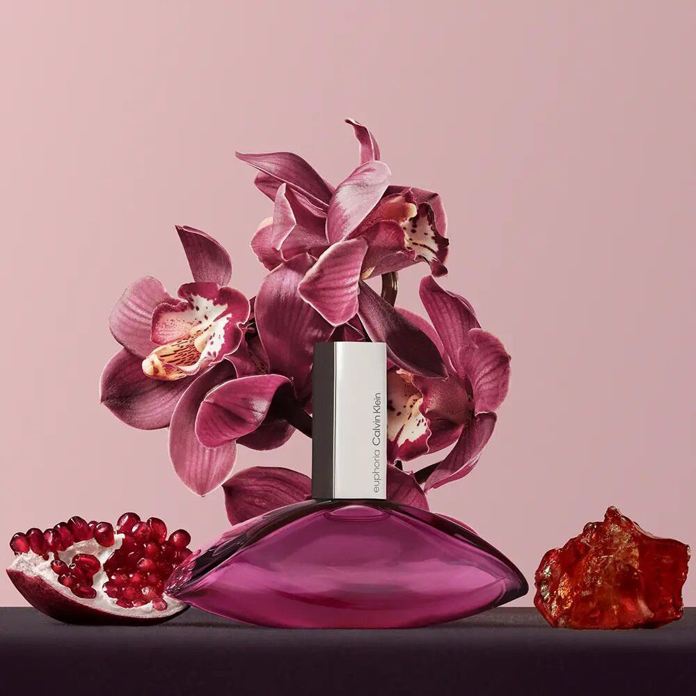 Euphoria-fles met zilveren dop, omringd door orchideeën, granaatappel en een amberkleurig object. Roze achtergrond.
