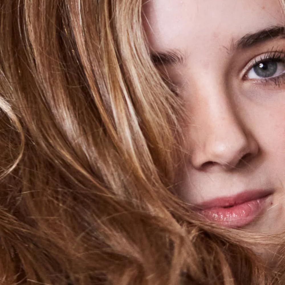 Portrait d'une femme avec des cheveux ondulés brun clair. Une partie du visage est visible, y compris l'œil et le nez.