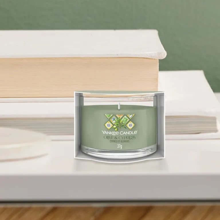 Bougie verte en verre et emballage. Inscription: Yankee Candle, Olive & Cypress, 37g. Sur une table avec des livres.