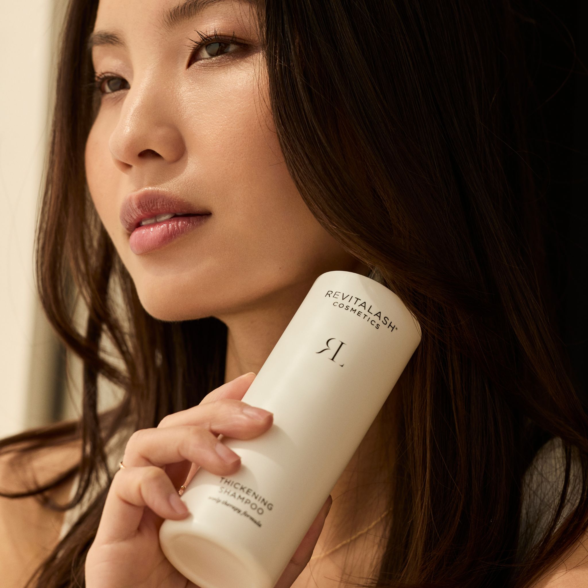 Femme aux cheveux longs tenant un flacon RevitaLash® Cosmetics Thickening Conditioner.