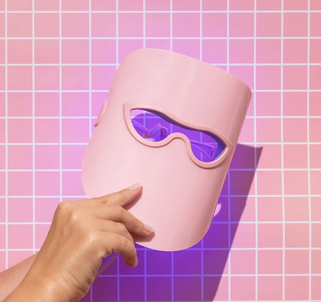 Un masque LED rose tenu dans une main devant un fond à carreaux.