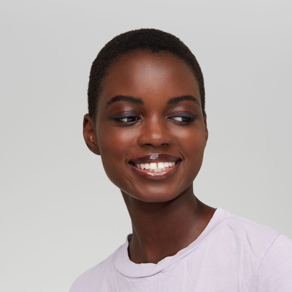 Portrait d'une femme souriante à la peau foncée. Elle porte un t-shirt clair et a les yeux maquillés.