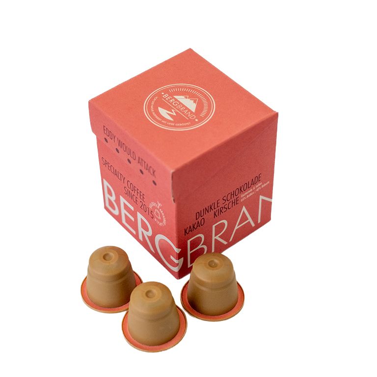 Trois capsules de café à côté d'une boîte rouge. Inscription : BERGBRAND, Eddy would Attack, Kapselkaffee.