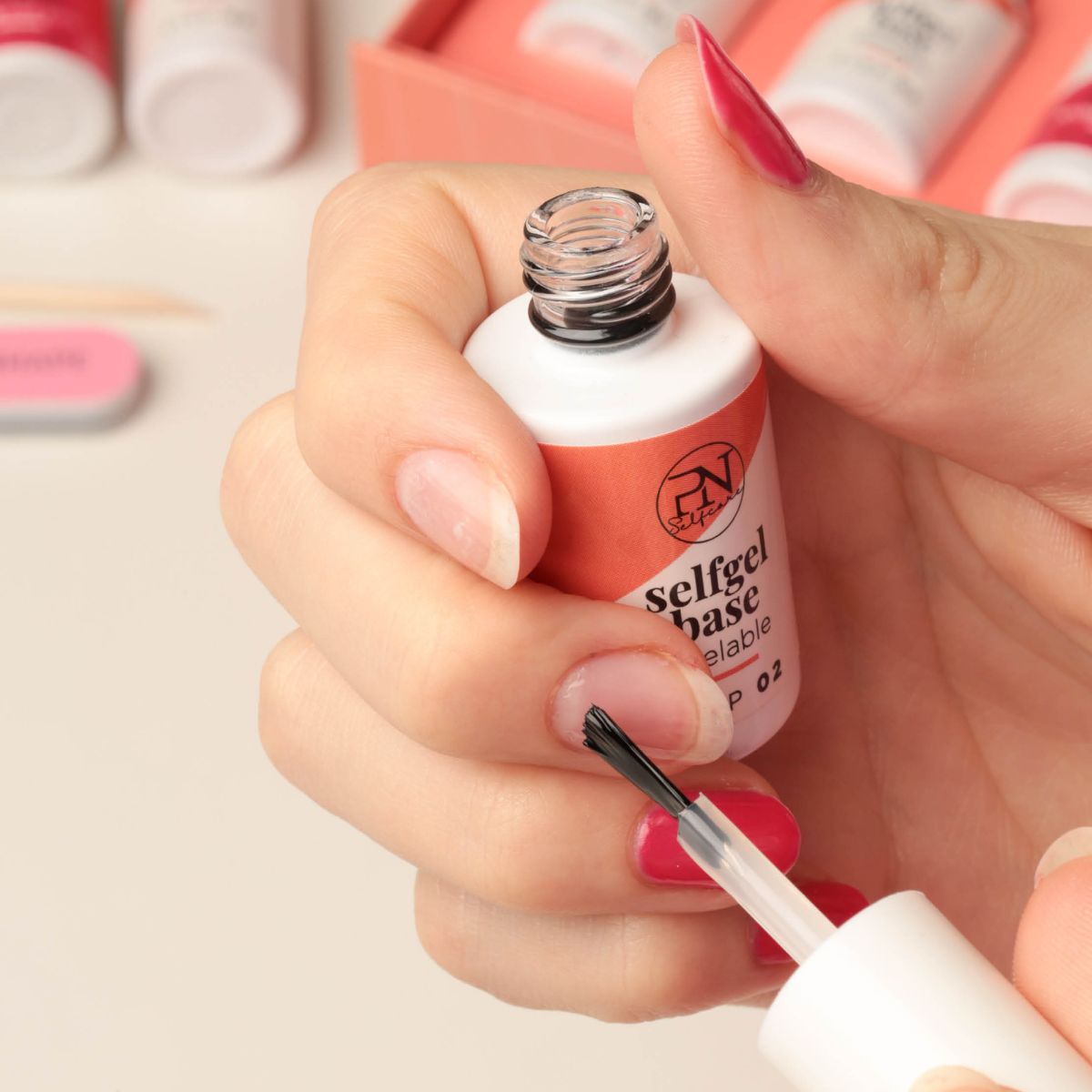 Hand houdt fles met borstel. Tekst: selfgel base peelable, STEP 02. PN-logo. Nagel met rode nagellak.
