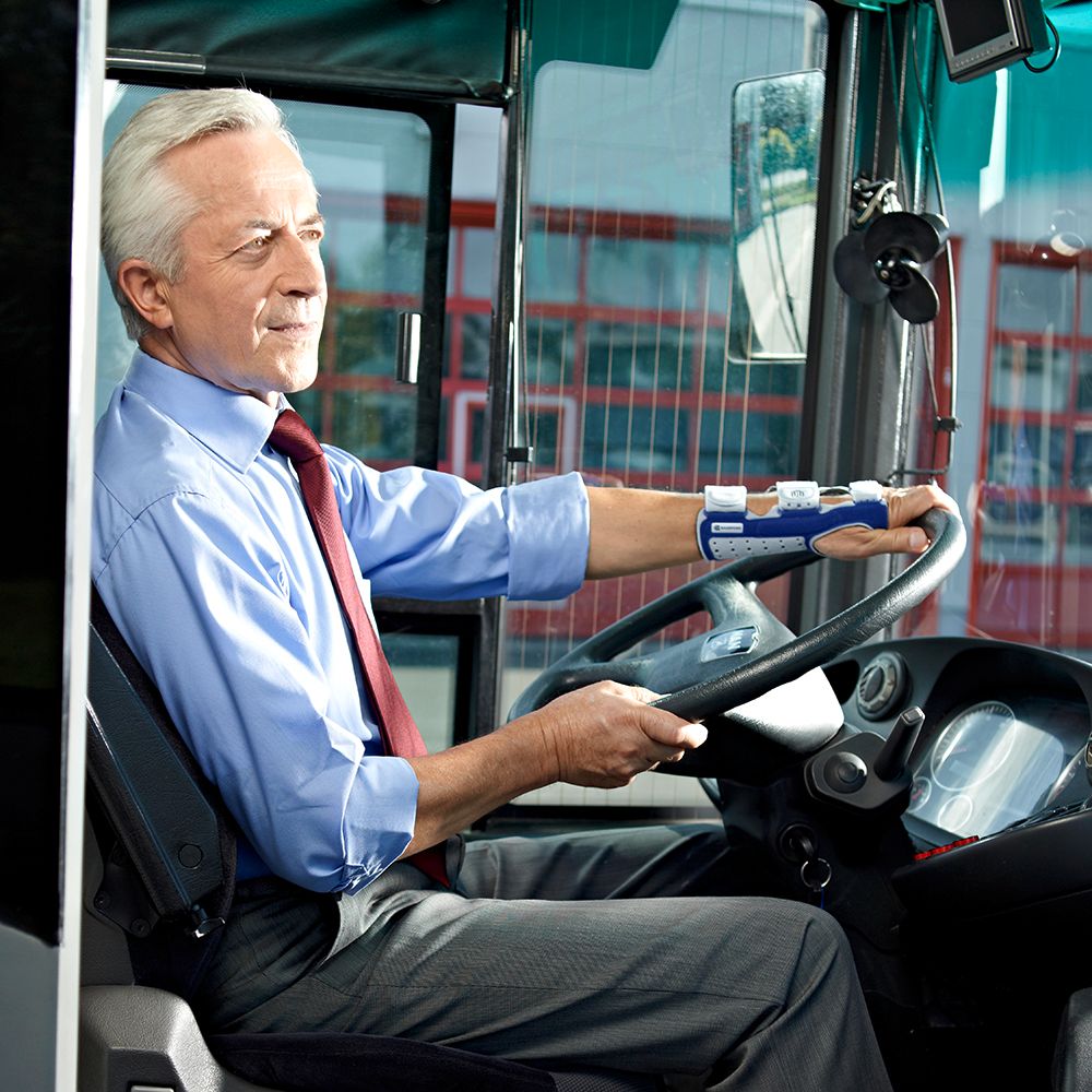 Man achter het stuur van een bus met een polsbrace. Hij draagt een overhemd en een stropdas.
