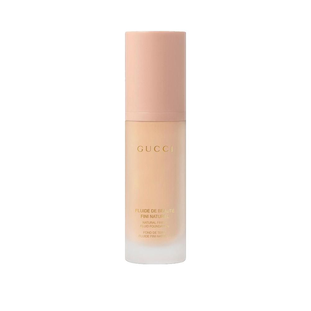 Gucci Fluide de Beauté. Flacon avec bouchon rose. Inscription Gucci. Liquide beige visible. Fond de teint.