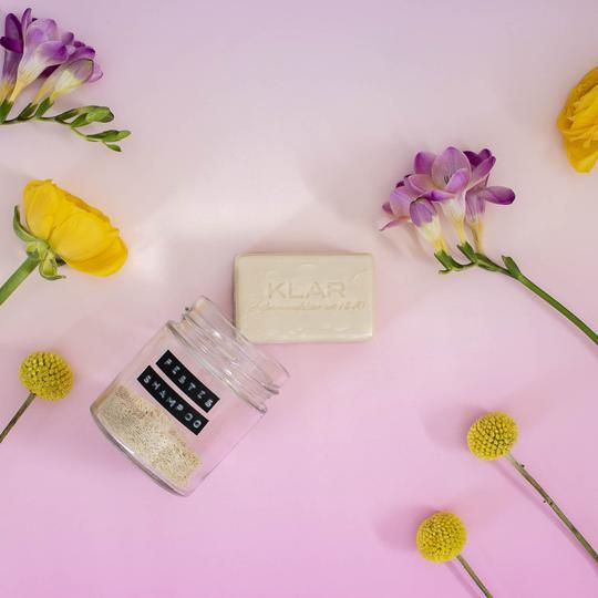 Vaste shampoo, glazen pot en bloemen op roze achtergrond. Shampoo rechthoekig. Pot met tekst: Vast Shampoo. Merk: Klar.