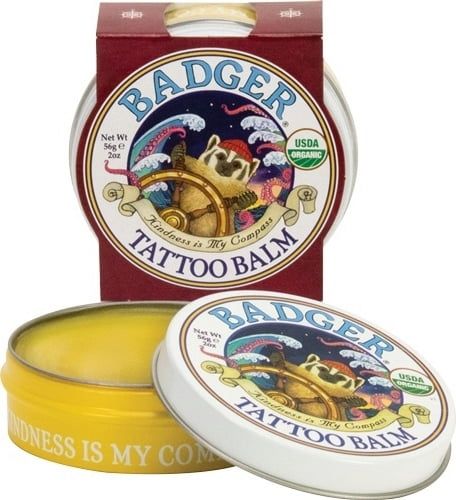 Badger Tattoo Balm. Ronde pot met deksel, gele inhoud en rode verpakking. Opschrift: BADGER, TATTOO BALM, USDA Organic.